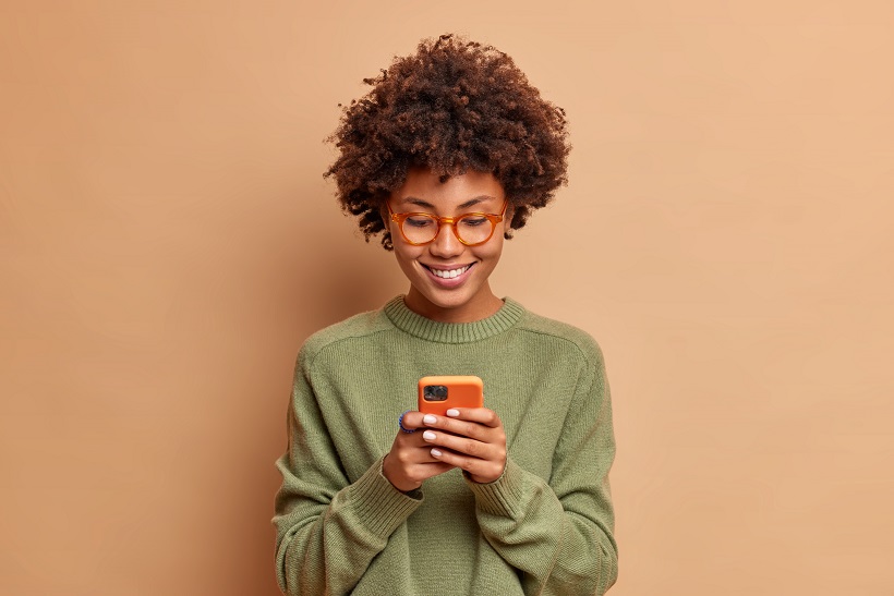 Isolated shot of African American woman uses smartphone application enjoys browsing social media creats news content makes online order wears spectacles and casual jumper poses over beige studio wall