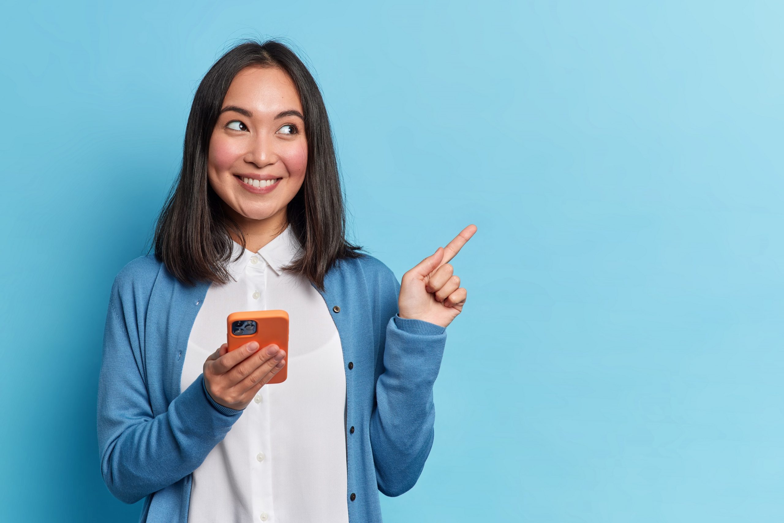 Beautiful Asian woman uses smartphone app sends messages in social media chat points away on copy space wears casual jumper isolated over blue background shows copy space for your advertisement